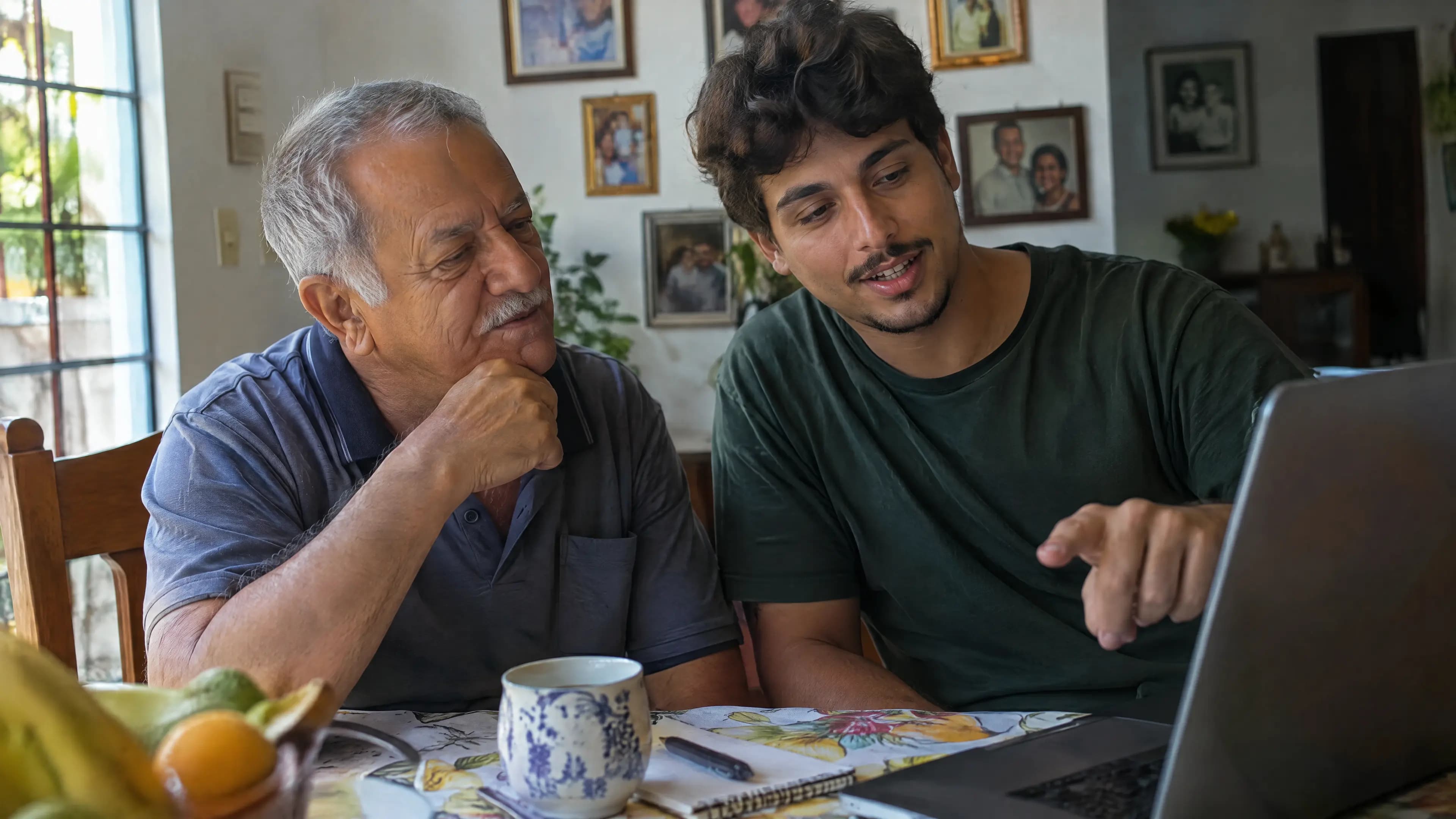 Neto mostra algo no notebook ao avô, que escuta com atenção, na mesa de jantar da família