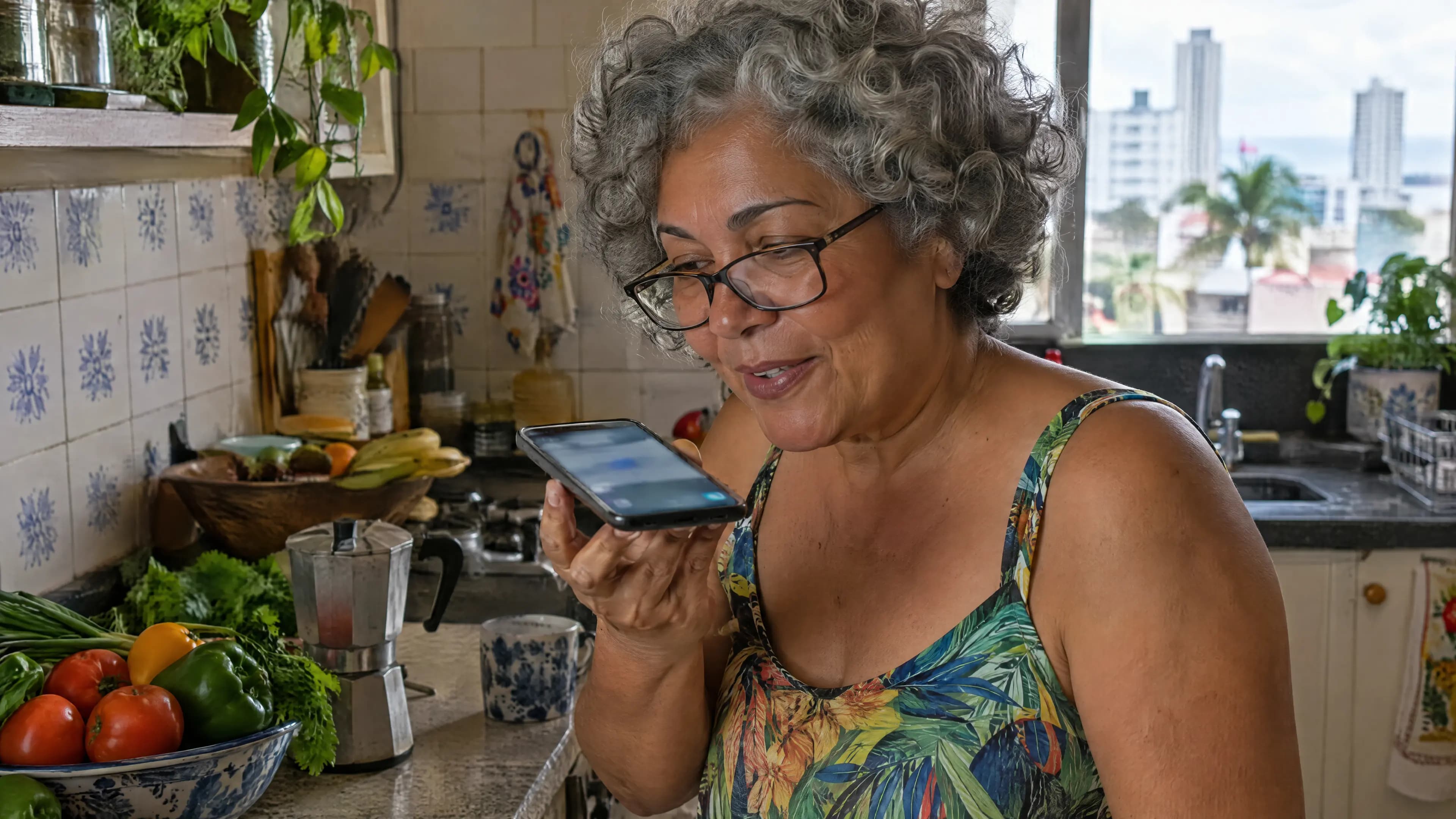 Mulher de cabelos grisalhos e óculos, na cozinha de casa, fazendo uma pergunta ao celular com um sorriso curioso