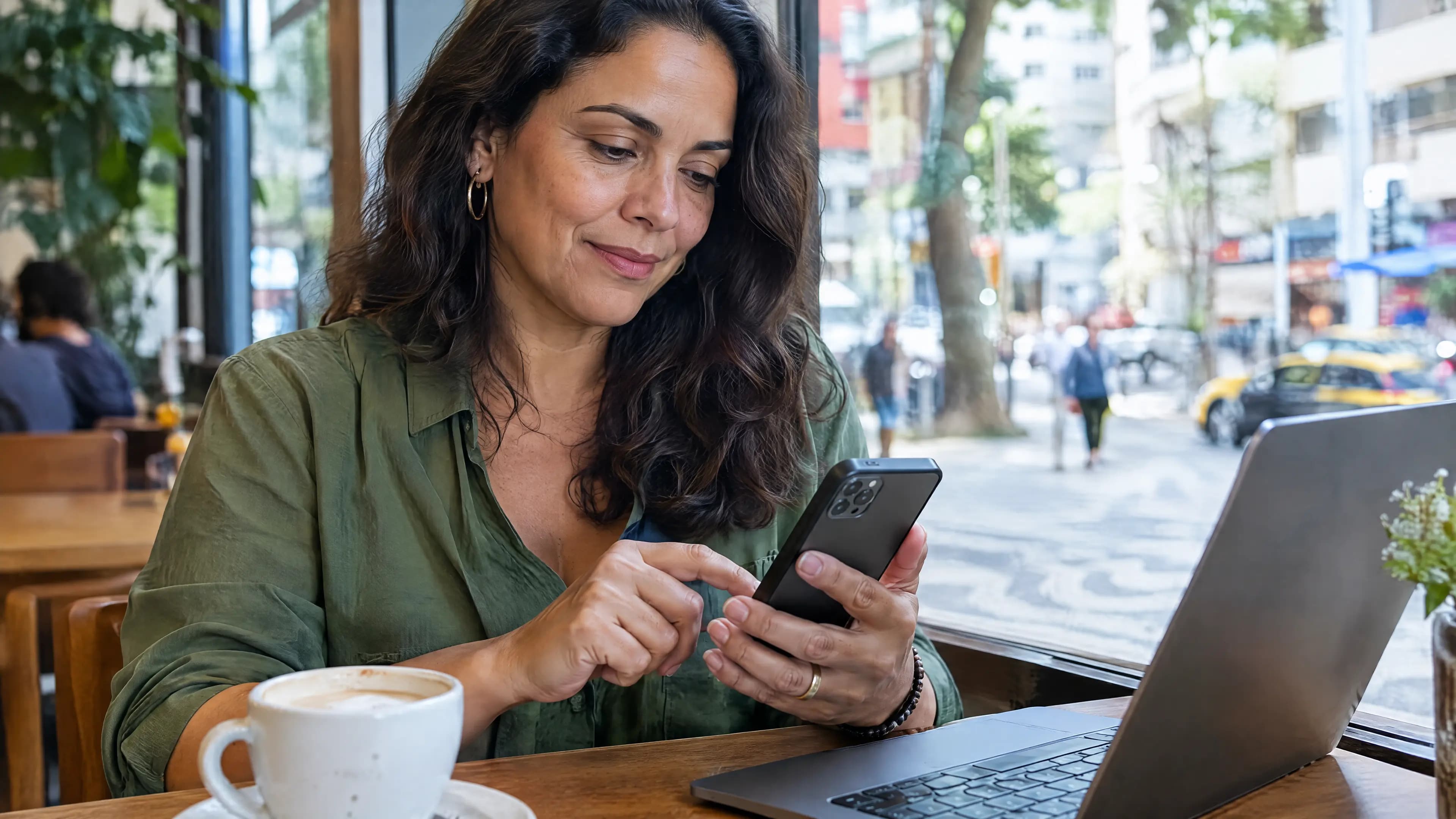 Mulher à mesa de um café, junto à janela, desbloqueia o celular com a digital, com o notebook e um café ao lado