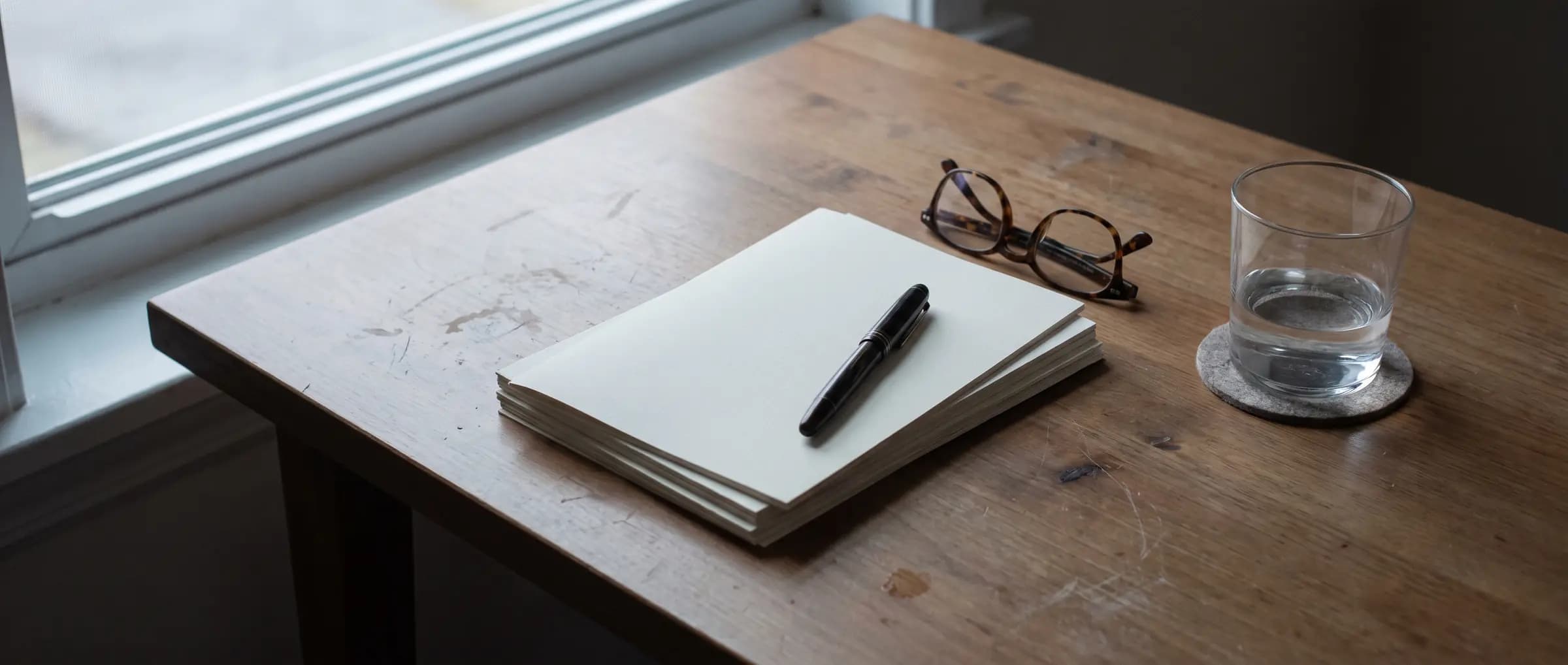 Pilha de folhas impressas com caneta-tinteiro, óculos de leitura e um copo d'água sobre uma mesa de madeira