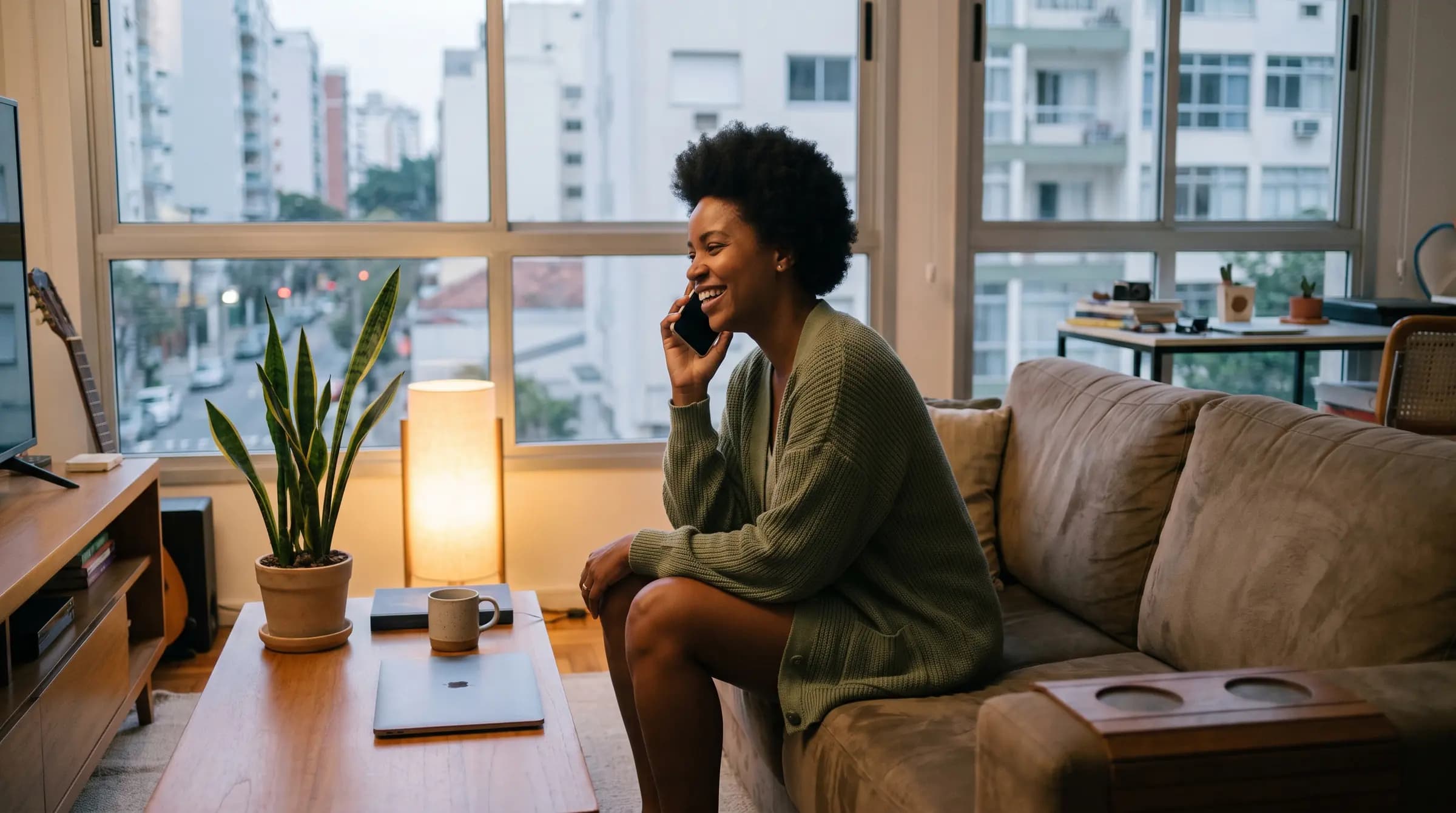 Mulher sentada no sofá de casa conversando pelo celular, com a cidade pela janela ao entardecer