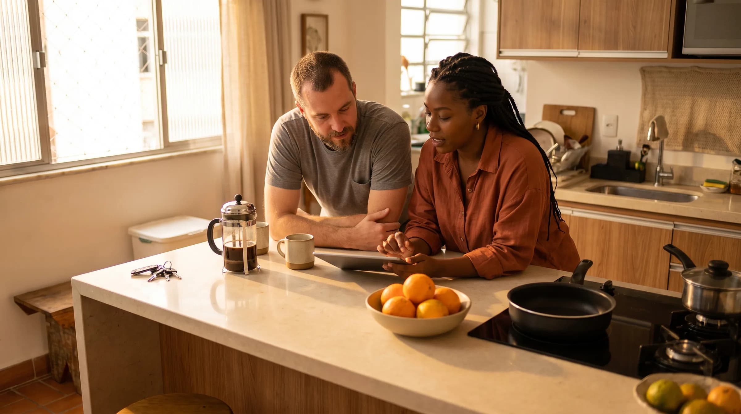 Casal conversando apoiado na bancada da cozinha, em um apartamento com luz da manhã