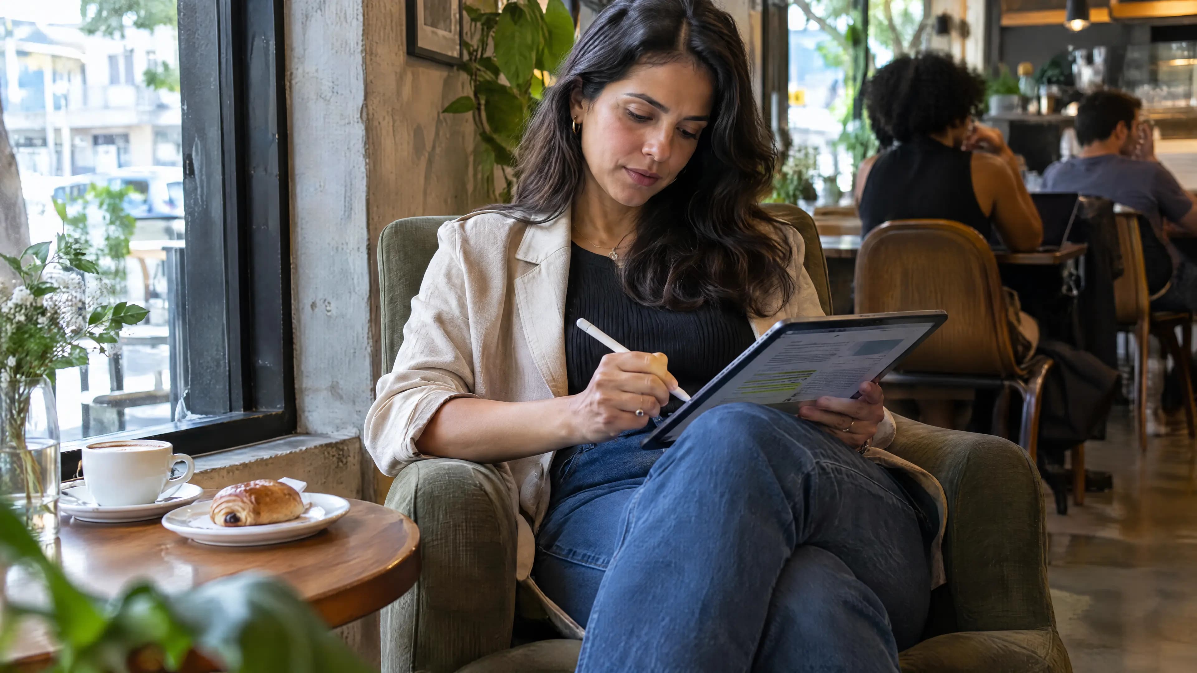 Mulher numa poltrona de café faz anotações no tablet com a caneta, com um café e um croissant na mesa ao lado