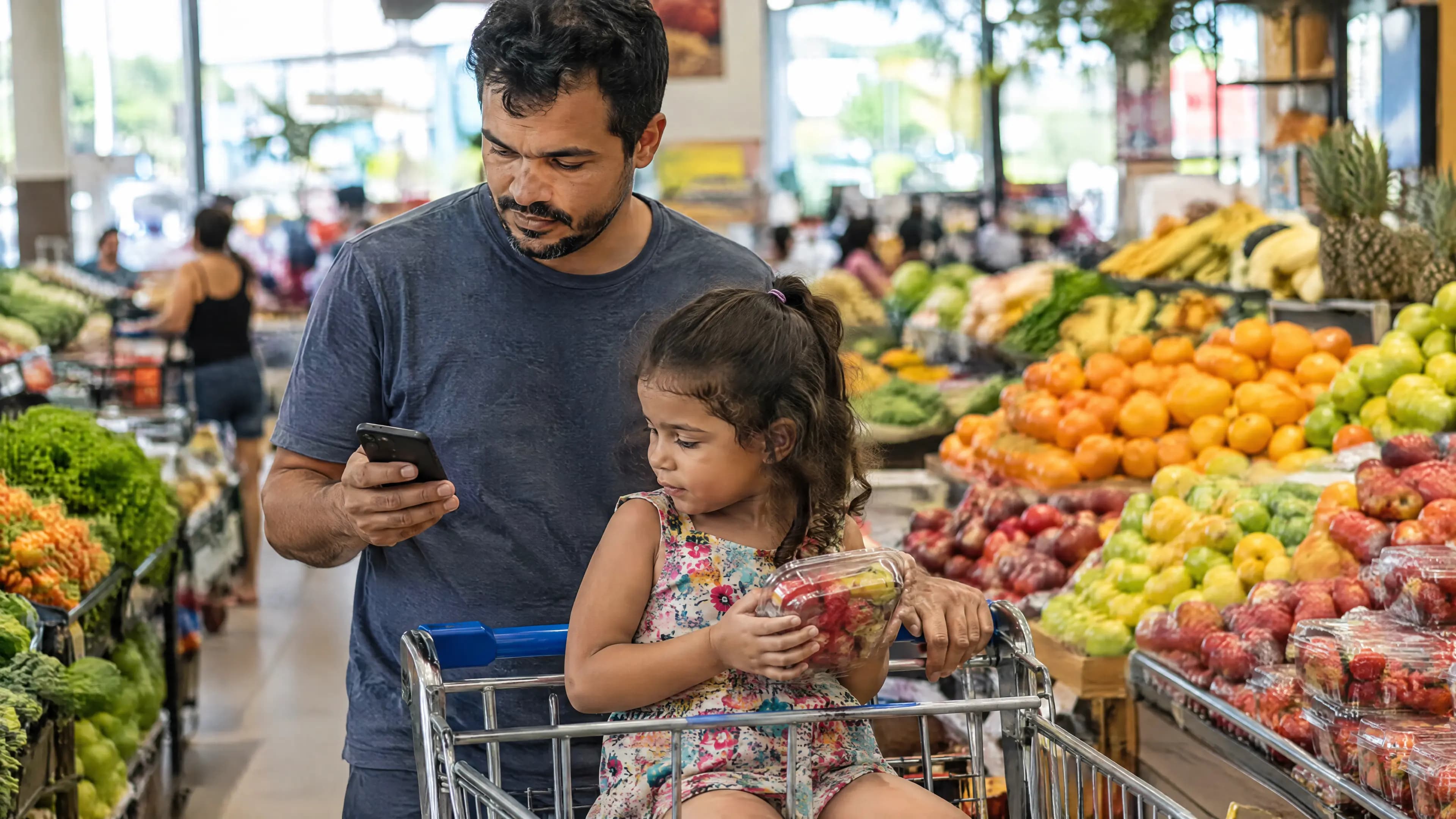 Pai empurra o carrinho no corredor de frutas do supermercado olhando a lista no celular, com a filha sentada no carrinho