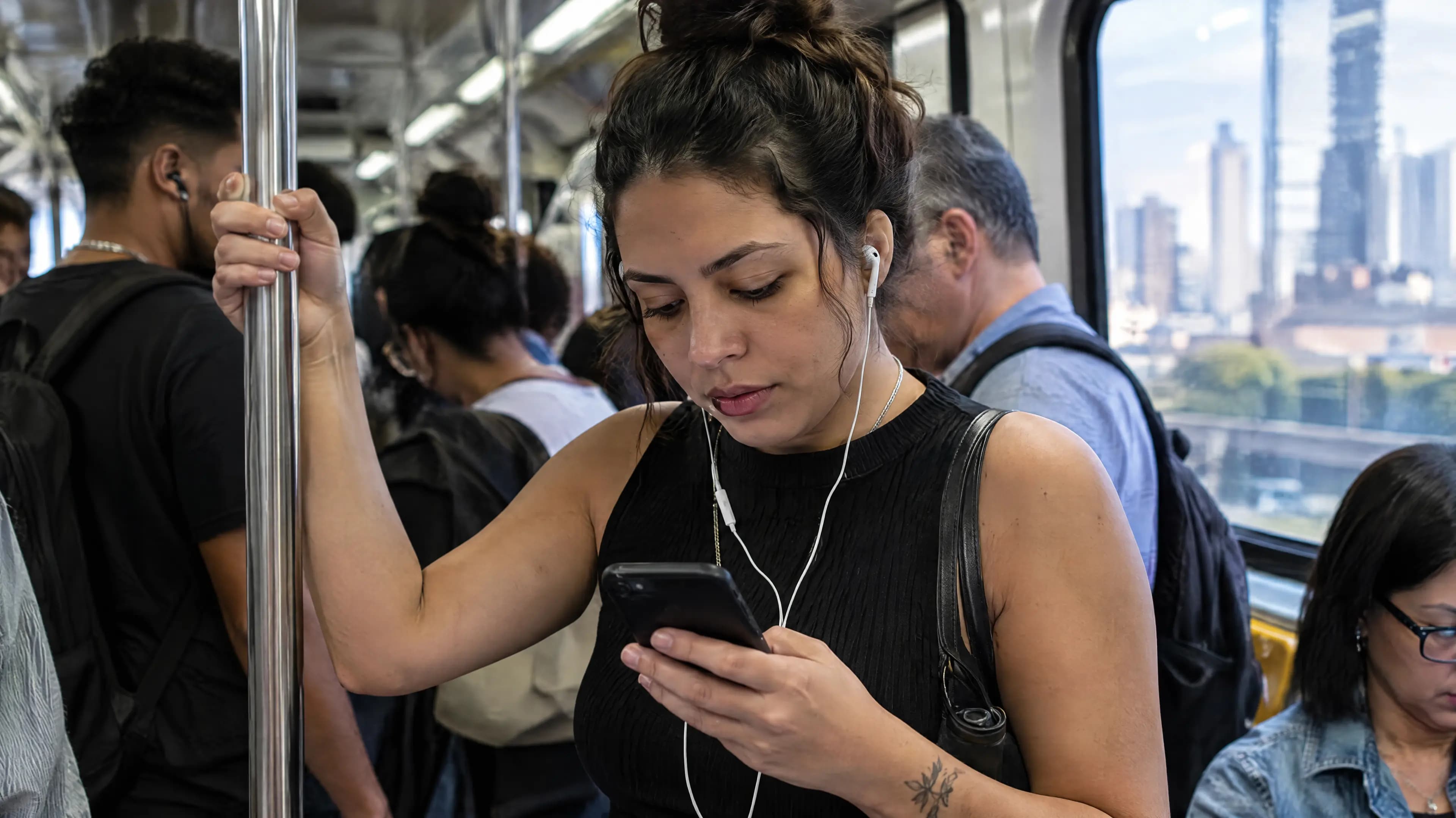 Moça em pé no vagão do metrô de manhã, segurando a barra e lendo notícias no celular, com fones de ouvido