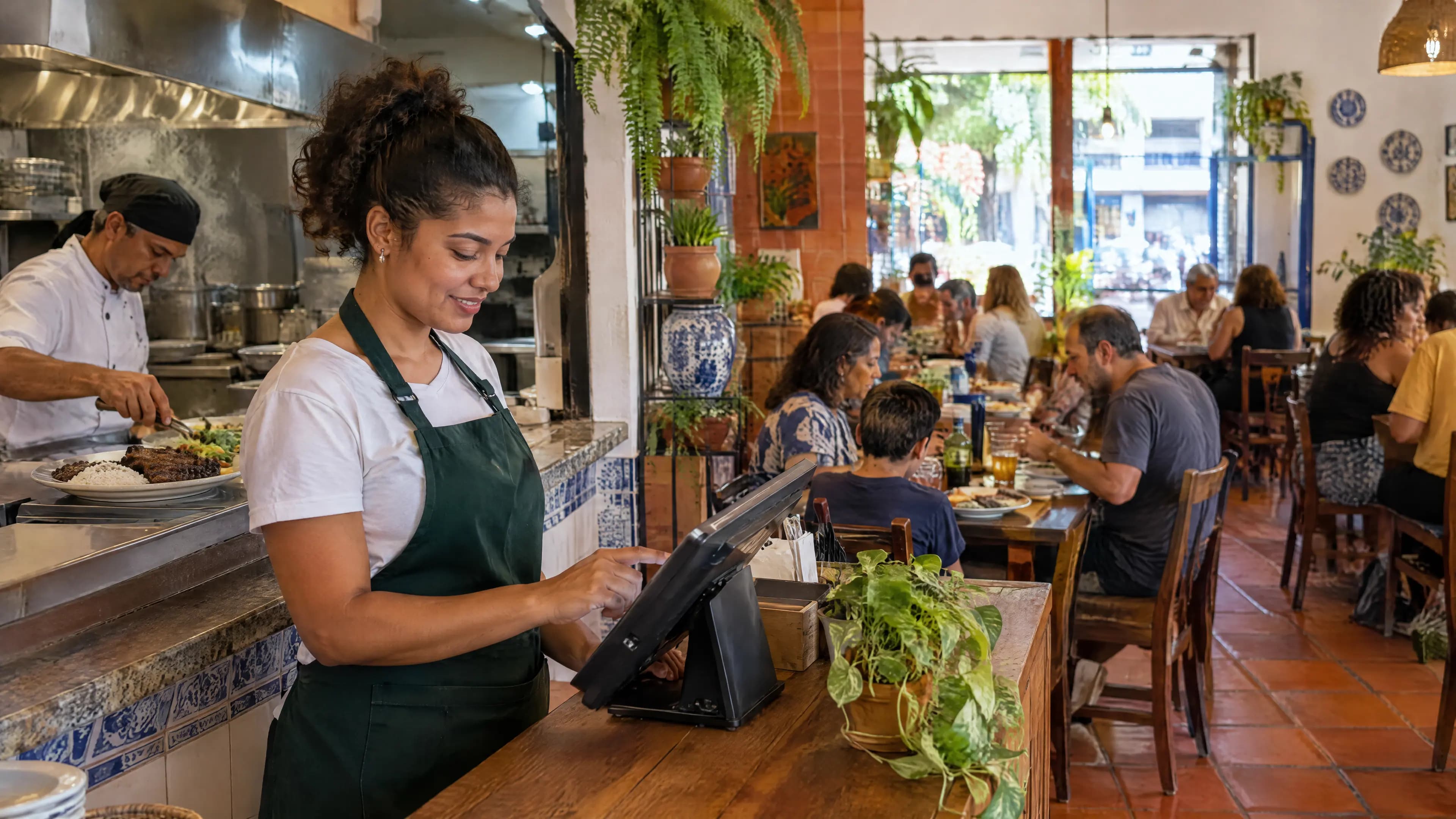 Atendente de avental verde lança o pedido no tablet do PDV no balcão, com o cozinheiro montando um prato de arroz, feijão e carne e o salão cheio ao fundo